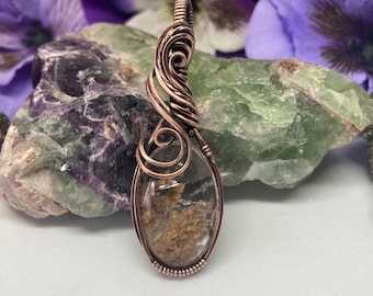 May include: A copper wire wrapped pendant featuring a clear quartz stone with brown and black inclusions. The pendant is oval shaped and has a delicate wire design.