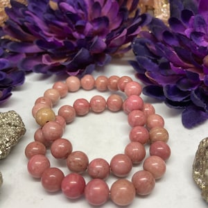 May include: Two pink gemstone bracelets made of round beads. The bracelets are stacked on top of each other and are lying on a white surface.
