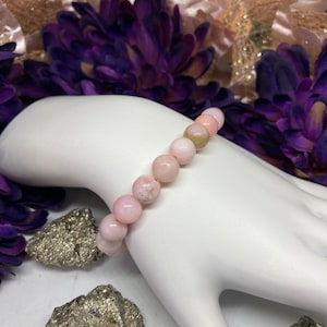 Könnte beinhalten: Ein rosafarbenes Opal-Perlenarmband mit einem hellrosa und weißen Farbschema. Das Armband besteht aus runden Perlen und wird auf einer weißen Mannequinhand gezeigt.
