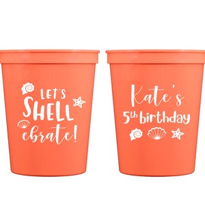 May include: Two orange plastic cups with white text. One cup says "Let's Shell-ebrate!" with a seashell and starfish design. The other cup says "Kate's 5th birthday" with a seashell and starfish design.