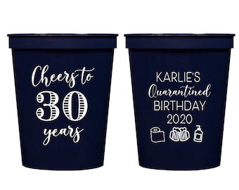 May include: Two blue plastic cups with white text. One cup says "Cheers to 30 years". The other cup says "Karlie's Quarantined Birthday 2020" with icons of toilet paper, beer, and hand sanitizer.