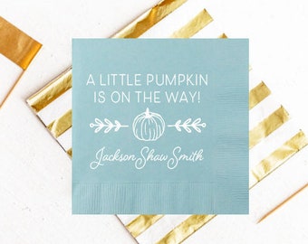 May include: A light blue baby shower napkin with gold stripes. The napkin has white text that reads "A little pumpkin is on the way!" with a small pumpkin graphic and the name "Jackson Shaw Smith".