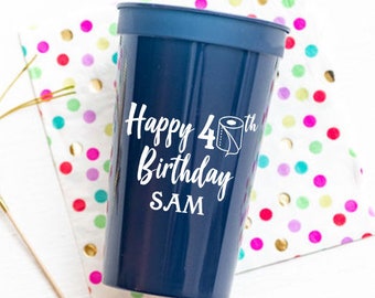 May include: A navy blue plastic cup with white text reading "Happy 4th Birthday SAM" and a toilet roll graphic. The cup rests on a polka dot napkin with gold and colourful dots.