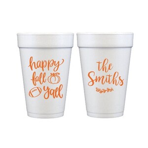 May include: Two white foam cups with orange lettering. One cup says "happy fall y'all" with a pumpkin and football graphic. The other cup says "the Smiths" with a leaf graphic. Perfect for fall gatherings.
