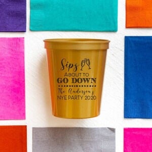 May include: Gold plastic cup with black text that reads "Sips About To Go Down The Anderson's NYE Party 2020".