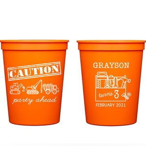 May include: Two orange plastic cups with white text. One cup says "CAUTION party ahead" with construction vehicle graphics. The other cup says "GRAYSON turns 3 FEBRUARY 2021" with a toolbox and tools graphic.