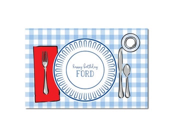 May include: A blue and white checkered placemat with a red napkin, a white plate with a blue rim, a fork, a knife, a spoon, and a small white plate. The plate has the text "happy birthday FORD" written on it.