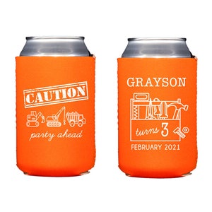 May include: Two orange and white insulated beverage can coolers. One cooler says "Caution party ahead" with a line of construction vehicles. The other cooler says "Grayson turns 3 February 2021" with a toolbox and tools.