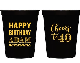May include: Two black plastic cups with gold lettering. One cup says "Happy Birthday Adam" and the other cup says "Cheers to 40".
