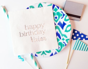 May include: White paper napkin with the text "happy birthday Vivian" in silver lettering. The napkin is on top of a blue and green patterned plate.