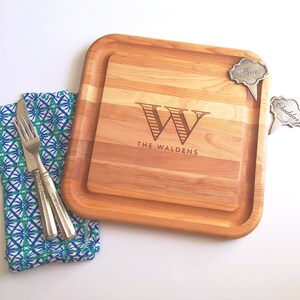 Pode incluir: Uma tábua de cortar de madeira personalizada com uma borda elevada. A tábua é gravada com a letra "W" e o texto "The Waldens". Inclui dois pequenos marcadores de queijo de metal, um com a etiqueta "Brie" e o outro com "Cheddar".