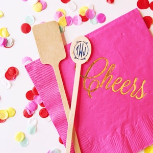 May include: Two wooden cocktail stirrers, one with a round, monogrammed topper and the other with a flat paddle-shaped topper. The stirrers are on a pink napkin with the word "Cheers" written in gold lettering.