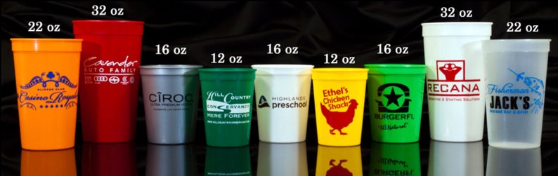 May include: A collection of ten plastic cups in various sizes and colors, including orange, red, silver, green, white, and yellow. The cups are printed with different logos and text, including "Casino Royale", "Cavendor Auto Family", "Ciroc", "Hill Country", "Highlands Preschool", "Ethel's Chicken Shack", "Burgerfl", "Recana", and "Fisherman's Jack's". The cups are arranged in a row from largest to smallest, with the largest cup being 32 ounces and the smallest cup being 12 ounces.