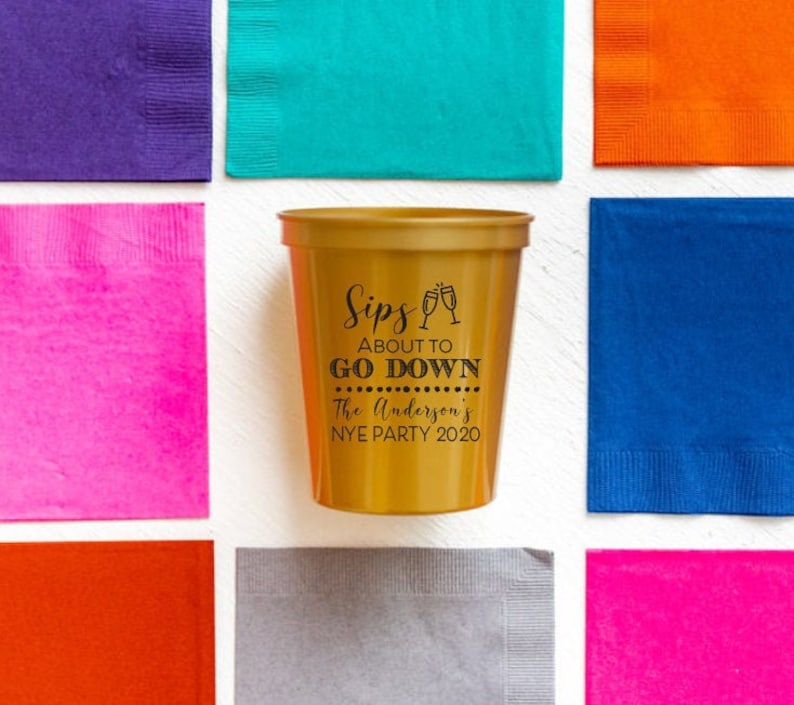 May include: Gold plastic cup with black text that reads "Sips About To Go Down The Anderson's NYE Party 2020".
