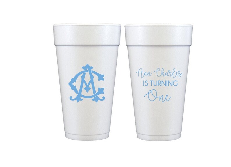 May include: Two white foam cups with blue text and designs. One cup says "Ann Charles is turning One" and the other has a blue monogram.
