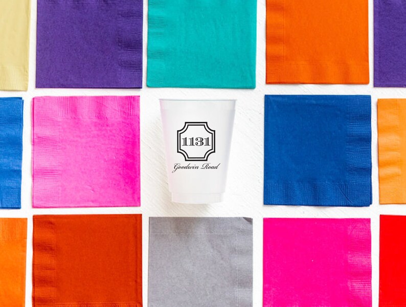 May include: A white disposable cup with a black border and the text "1131 Goodwin Road" printed on it. The cup is surrounded by colorful napkins in shades of pink, blue, orange, and purple.