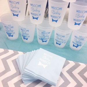 May include: A stack of blue paper napkins with the text "Welcome Baby Wilkerson" and a blue bow tie printed on them. There are also several clear plastic cups with the same text and bow tie design printed on them.