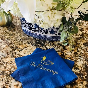 May include: Two blue napkins with gold embroidery. The top napkin features a gold duck and the text "The Mannings".