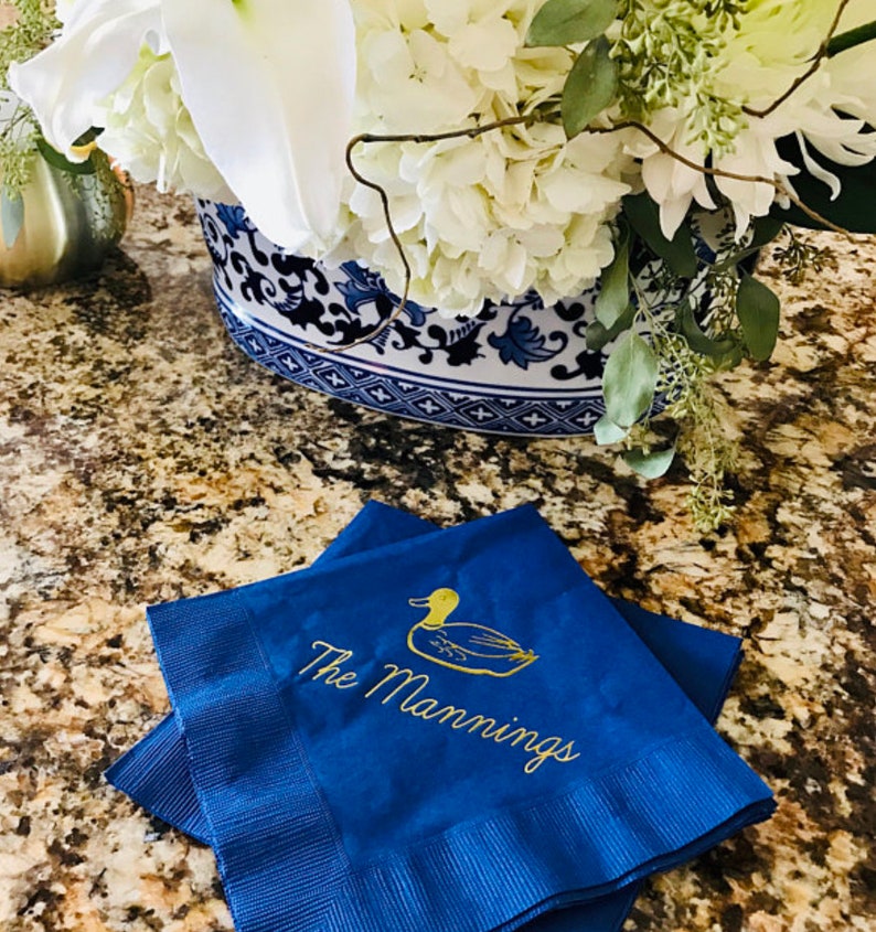 May include: Two blue napkins with gold embroidery. The top napkin features a gold duck and the text "The Mannings".