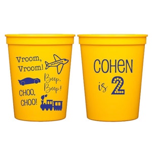 May include: Two yellow plastic cups with blue text. One cup says "Vroom, Vroom!" with a plane, car, and train. The other cup says "COHEN is 2" with a number 2.