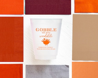 Könnte beinhalten: Ein weißer Plastikbecher mit dem Text "GOBBLE till you wobble" in Orange und einer Truthahn-Grafik. Der Becher ist von orangefarbenen, braunen und weinroten Servietten umgeben, ideal für eine Thanksgiving-Feier.