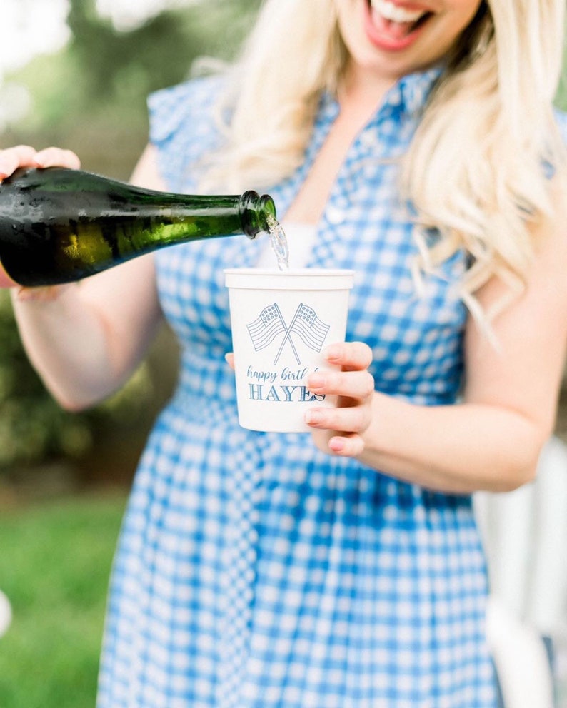 May include: A person pouring champagne into a white paper cup with a blue and white American flag design and the text "happy birthday HAYES".
