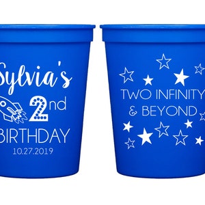 May include: Two blue plastic cups with white text. One cup says "Sylvia's 2nd Birthday 10.27.2019" with a rocket ship graphic. The other cup says "Two Infinity & Beyond" with stars.