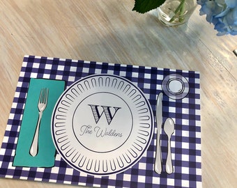 May include: A blue and white gingham placemat with a white plate featuring a black 'W' and the text 'The Waldens'. The placemat also includes a fork, knife, spoon, and a small white plate.