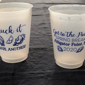 May include: Two white plastic cups with blue text and images. The first cup says "Shuck it" with an oyster and a lemon wedge. The second cup says "Get to The Point Spring Break Alligator Point, FL 2020" with two oysters.