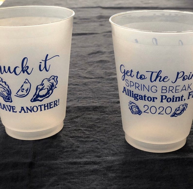 May include: Two white plastic cups with blue text and images. The first cup says "Shuck it" with an oyster and a lemon wedge. The second cup says "Get to The Point Spring Break Alligator Point, FL 2020" with two oysters.