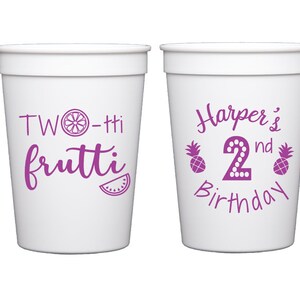 May include: Two white plastic cups with purple text. One cup says "TWO-tti frutti" with a lemon and watermelon slice. The other cup says "Harper's 2nd Birthday" with two pineapples.