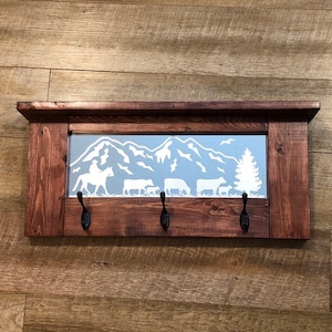 May include: A rustic wooden coat rack with a mirror featuring a white silhouette of a cowboy riding a horse and cattle in a mountain landscape. The rack has three black metal hooks.