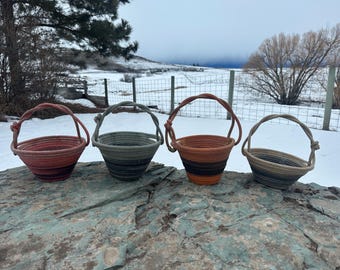 May include: Four small rope baskets with handles, in shades of red, gray, and brown, sitting on a gray rock surface. The baskets are made of woven rope and have a rustic, handmade look.
