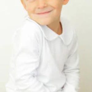 May include: A young child wearing a white long-sleeved shirt with a rounded collar and orange and white checkered pants. The child is smiling and looking towards the camera.