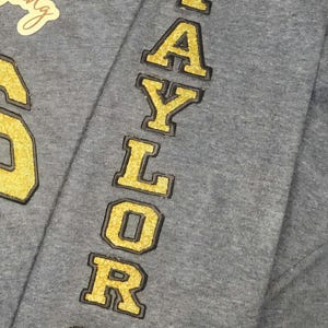 May include: Close-up of a gray sweatshirt sleeve with the name "TAYLOR" in gold glitter lettering down the side. The number "26" is also in gold glitter. The word "Bling" is visible in gold script.