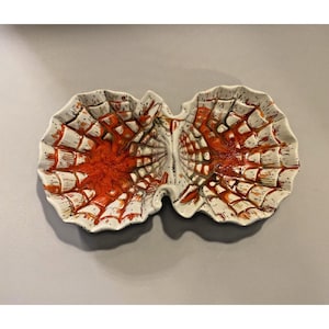 May include: A ceramic serving dish with a unique design. The dish is divided into two sections, each shaped like a leaf. The interior is a vibrant orange, with a white and orange web-like pattern. The dish is speckled with black dots.