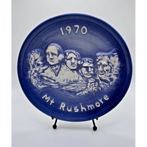 May include: A round, deep blue ceramic decorative plate featuring a white embossed relief of the Mount Rushmore monument. The text 1970 is printed at the top, and the text Mt Rushmore is printed at the bottom in white, raised lettering.