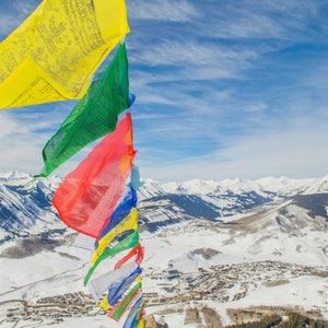 May include: Colorful prayer flags flutter in the wind against a backdrop of snow-covered mountains and a bright blue sky. The flags are yellow, green, red, and blue, adding a vibrant contrast to the winter landscape.