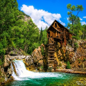 May include: A wooden water mill with a water wheel sits on a rocky cliff next to a waterfall. The mill is surrounded by lush green trees and a blue sky with white clouds.