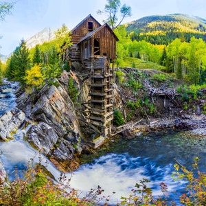 May include: A wooden water wheel mill in a mountainous landscape with a rushing river and colorful fall foliage. The mill is built into the side of a rocky cliff.