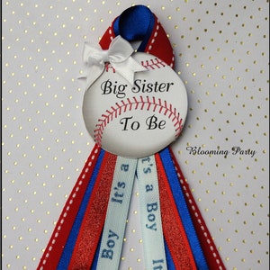 May include: A baseball-themed baby shower ribbon with the text "Big Sister To Be." The ribbon features red, white, and blue colors, with the words "It's a Boy" on a light blue ribbon. A white bow is at the top.