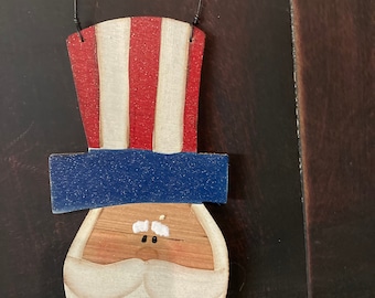 May include: A wooden ornament shaped like a face with a red, white, and blue top hat. The face has a white beard, black eyes, and a brown nose.