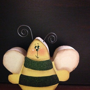 May include: A wooden bee figurine with yellow and black stripes, white wings, and black antennae. The bee has a painted face with black eyes and a red mouth.