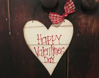 May include: A heart-shaped wooden sign with the words "Happy Valentines Day" in red script. The sign is cream-colored with horizontal wood grain lines. A red and white gingham bow is attached at the top. The sign hangs from a black wire.