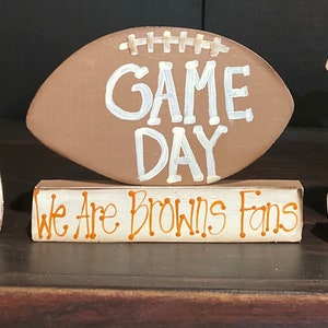 May include: A brown wooden football with white stitching and the words "GAME DAY" painted on it. A smaller wooden block with the words "We Are Browns Fans" painted in orange on it sits below the football.