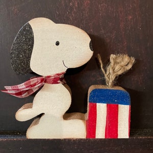 May include: A white wooden figurine of a dog wearing a red and white checkered scarf, sitting next to a red, white, and blue wooden box with a burlap string.