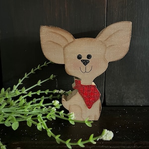 May include: A wooden figurine of a small dog with large ears, painted in a light brown color. The dog is wearing a red and black plaid bandana.