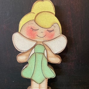 May include: A wooden figurine of a fairy with closed eyes, blonde hair, a green dress, and brown wings. The fairy is painted with blush on her cheeks.