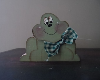 Peut inclure: Une figurine de grenouille en bois vert avec un sourire peint en blanc et des yeux noirs. La grenouille porte un nœud papillon à carreaux verts et blancs.
