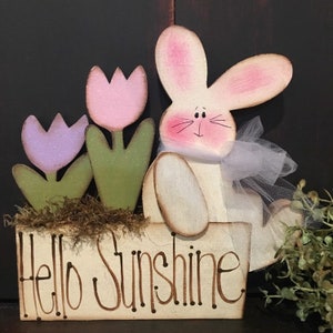 以下が含まれることがあります： 「Hello Sunshine」と茶色で書かれた木製の看板。ピンクの耳と薄い紫色のチュールリボンをつけた白いウサギが看板の上に座っている。ウサギの後ろには、ピンク、紫、緑の3つの木製のチューリップがある。